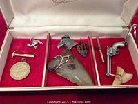Open treasure box showing shark teeth wired for necklace, several metal and wood charms, and a gun-shaped keychain inside a red velvet-lined box.