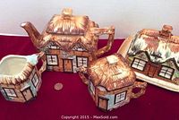 Tea set items arranged on red cloth with a penny for size comparison. Includes teapot, creamer, sugar bowl, and covered dish. Each piece shaped like cottages with distinctive thatched roofing and handpainted details.