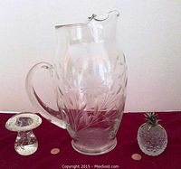Photo showing large glass pitcher with floral etching and metal rim, solid glass toadstool and pineapple figurines with pennies for size comparison