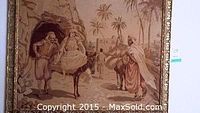 Framed tapestry hanging on a wall showing a detailed scene with several figures, camels, palm trees, and village structures. The frame is ornate and gold-colored.
