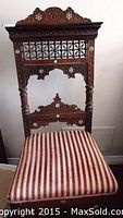 Front view of an ornate carved wooden chair with patterned inlay on the backrest and an upholstered seat with red and white stripes.