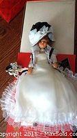Bride doll wearing a white dress with lace trims and large black and white hat, displayed lying with box background.