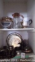 Shelf showing a variety of silver plated trays, a glass teapot with brass handle and lid, cream and sugar set, and brass finish cups.