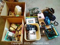 Overview of multiple boxes and bins containing various electronics, cameras, phones, accessories, and foreign currency.