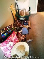 Cluster of assorted household items including laundry basket, pillow, bags, wrapping paper, towels, and straw hat laid on floor near wall.
