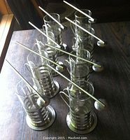 Full set of 8 tall clear glass bar glasses with steel metal holders, metal coasters, and long spoons with hollow handles used as straws, arranged on wooden table.