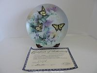 Front view of the limited edition plate showing the butterfly and floral design on a pastel background, displayed on a wooden plate stand alongside the certificate of authenticity.