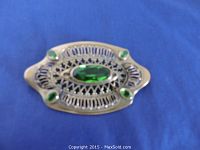 Photo of a Victorian-style brooch with a large central green brilliant cut crystal and four smaller green crystals in an intricate silver-tone metal setting, placed on a blue background.