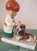 Side view of porcelain figurine showing boy with red hair in white doctor coat holding medicine bottle and red piece, dog lying in front.