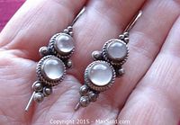 Front view of both earrings held in a hand showing two round bezel-set moonstones and decorative silver beads and twisted silver frames.