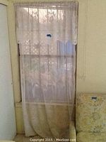 Full length view of one lace panel curtain hanging in front of a window and radiator, showing sheer fabric and lace pattern