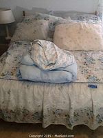 Photo showing full size bed with headboard, floral linens including bedspread, pillow shams and bed skirt, light blue comforter, and lamp on side table.