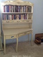 Full view of off-white vintage corner desk with two bookshelves filled with books, three small drawers below shelves and one larger drawer below desktop. Curved legs and intricate molding details.