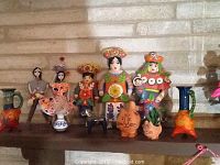 Full view of Mexican decorative figurines including colorful human characters, animal figurines, and ceramic vessels on a shelf against a stone wall.