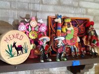 Photo showing all items on shelf including basket, banners, framed cloth art, and various figurines including skeleton musicians and tradition dressed figures.