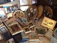 Photo showing assorted painted artworks, framed oval floral prints, and decorative mirrors all leaning against a wall.