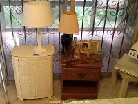 View showing white wicker hamper with ceramic lamp on top, wooden nightstand with dark ceramic lamp, and assorted decorative items on nightstand