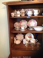 Full view of assorted china pieces displayed on left side of wooden hutch shelf including teapots, creamer, sugar bowl, cups, saucers, plates, bowl, and decorative items