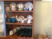 Wide view of shelving unit with assorted china plates, bowls, cups, and various glasses including blue glasses and clear stemmed glasses.