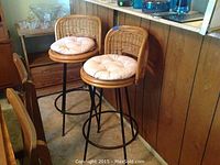 Two wicker swivel bar stools with removable white cushions, black metal swivel bases, positioned near wood-paneled counter.