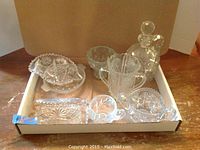 Assorted crystal/glass dishes shown in a cardboard box on a wooden table. Pieces include a two-handled ice bucket, covered candy dish, small decanter, creamer, and trays.