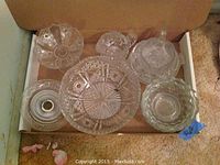 Photo showing assorted crystal and glass dish pieces in a box, varying shapes and sizes, some with intricate cut designs and one with a lid.