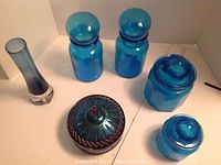 Photo of six blue glass items including two apothecary jars with round lids, one hexagonal jar with lid, candy dish with lid, and a tall vase.
