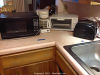 Photo shows four kitchen appliances on a countertop: a black microwave, white coffee maker, white toaster oven, and black toaster with cover near a sink.