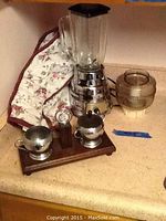 Electric blender with glass jar, chrome base, brown grinder attachment, wooden tray with sterling silver-plated small dishes, and an electric grinder attachment.