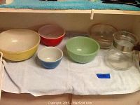 Photo of six items including four Pyrex mixing bowls in yellow, green, red, and blue, a clear glass coffee pot with silver band, and a clear glass lid or small bowl, all set on a white towel.