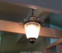 Side and slightly angled view of the hanging light fixture showing the glass shade glowing lit, metal starburst frame around top, ribbed metal bottom cap and metal chain.