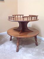 Full view of a wooden Hitchcock 2-tiered round corner table with turned legs and spindle railing around the upper tier, placed in a corner against two beige walls and carpeted floor.