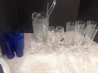 Photo showing large diamond pattern textured glass pitcher with ladle, blue tall drinking glasses, floral patterned crystal glasses, clear champagne flutes, and two small clear round items.