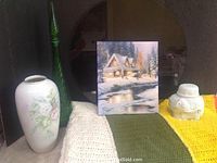 Photo showing Thomas Kinkade print of cottage in snow, crocheted afghan with three colors, two green glass vases, and white ceramic elephant figurine placed on a surface.