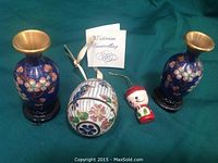 Photo showing two blue Victorian enamelled mini vases with pink flower patterns on black bases, a white floral cloisonne hanging ornament with gold accents, and a small painted wooden Santa head ornament with red hat and green collar, all displayed on a green fabric background.