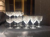 Photo shows 8 small shallow bowl clear cut glasses on the left and 7 larger round bowl clear cut glasses on the right, all with stems, placed on a black cloth.