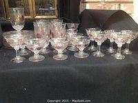 Photo showing a set of clear glassware and saucers, all decorated with painted purple grape clusters and gold leaf accents. Includes a variety of glass styles such as tall glasses and coupe glasses.