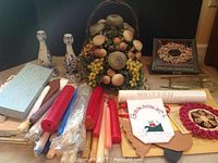 Photo showing assorted candles, two white and blue ceramic candle holders, silver wreath centerpiece decorated with faux grapes and round ornaments, holiday linens box, and rolled linens.