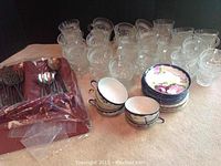 Photo showing 38 clear glass cups, 4 ceramic tea cups with floral saucers, and metal silverware in plastic sleeve on a tabletop.