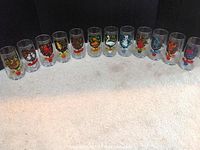 Photo showing the full set of 12 clear drinking glasses aligned in a row on carpeted floor. Each glass features colorful holiday illustrations and song verses related to the "12 Days of Christmas."