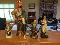 Four Chinese figurines displayed on a table including Guan Yu in green robes, Jiang Taigong fishing, and two men playing stringed instruments.
