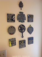 Photo showing 10 metal wall plaques arranged in 3 rows on a beige wall. Plaques have various kitchen and home sayings with decorative elements and black background with white/yellow text.