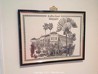 Framed black and white drawing of Raffles Hotel Singapore hanging on wall, showing detailed depiction of the hotel and surrounding palm trees within decorative border.