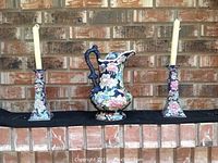 Photo showing the matching set of a floral patterned ceramic pitcher and two candlesticks with cream-colored candles.