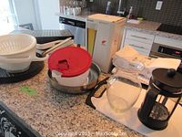 Photo showing assortment of kitchen items including salad spinner, frying pan, Bodum coffee maker, glass pitcher, place mats, and boxed glassware on countertop.