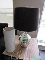 Chrome table lamp with black shade on wooden surface, white ceramic cylinder next to it