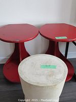 Pair of round distressed red wooden end tables on wheels accompanied by a natural wood cylindrical stool placed on the floor against a white wall.