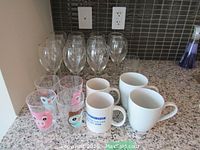 Photo showing an assortment of 4 large clear wine glasses, 5 small clear drinking glasses with cartoon faces, and 3 ceramic mugs on a kitchen counter.