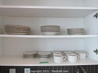 Wide shot showing stacks of dinner plates, salad plates, bread plates, and cups arranged on two shelves in a white cabinet.