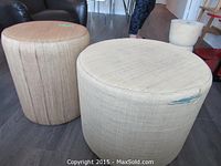 Two round fabric-upholstered ottomans shown on grey floor; larger beige one with tear visible and smaller brown one beside it.